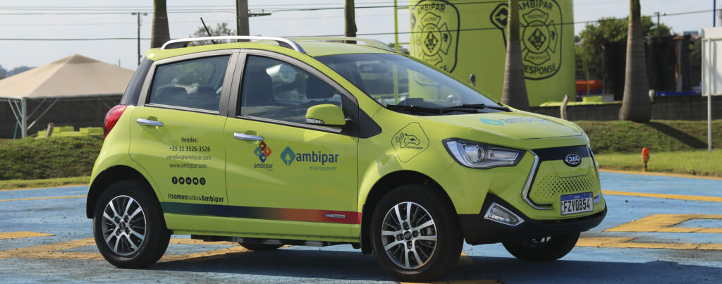 Veículo sustentável, de cor amarela, com inscrições nas portas. O carro está em um estacionamento a céu aberto, com piso azul e árvores altas no plano de fundo
