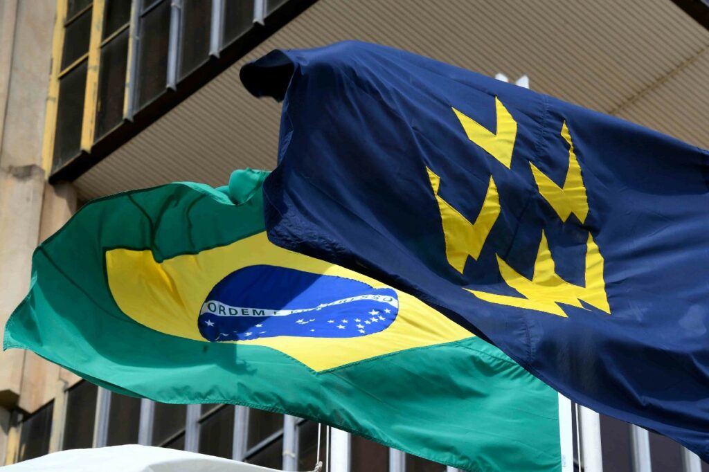 A imagem mostra duas bandeiras hasteadas em frente a um prédio. A bandeira à esquerda é a bandeira do Brasil e a bandeira à direita é a bandeira da CNI. Isso porque, o presidente, Ricardo Alban, analisa como a decisão do Banco Central afeta a indústria 