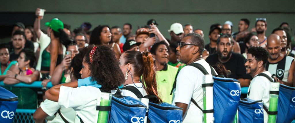Na imagem aparecem representantes da ação sustentável de reciclagem da Água do Rio durante o carnaval. Eles levam nas costas uma espécie de mochila azul, com a logo da marca. 