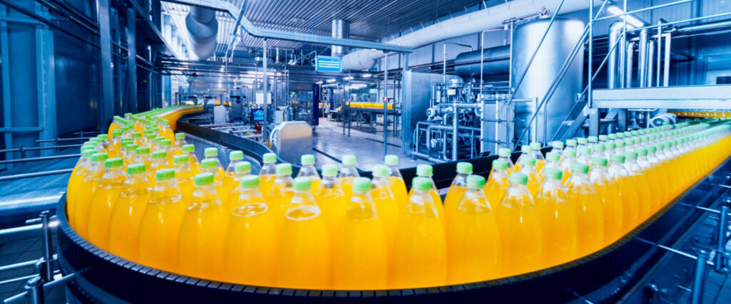 Na imagem aparecem garrafas plásticas transparentes, contendo um líquido amarelo. Elas estão em uma fileira sobre uma máquina em uma indústria