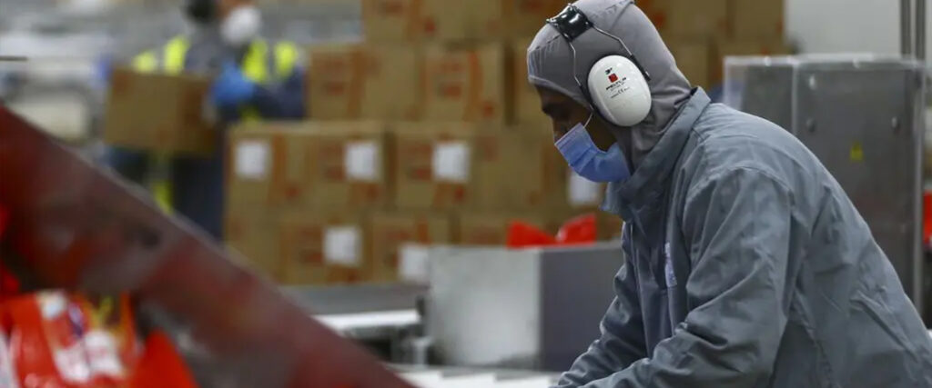 Trabalhador industrial realizando atividades, ele veste um uniforme de proteção de cor cinza. Usa máscara sobre o nariz e a boca e abafadores de sons nos ouvidos