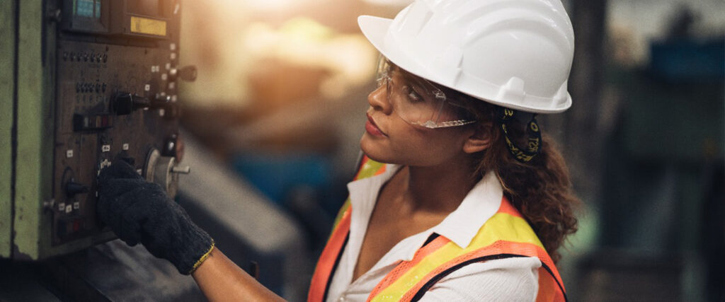 Imagem de uma mulher trabalhando na indústria. Ela usa um capacete e óculos de proteção, tem a pele negra e cabelos castanhos 